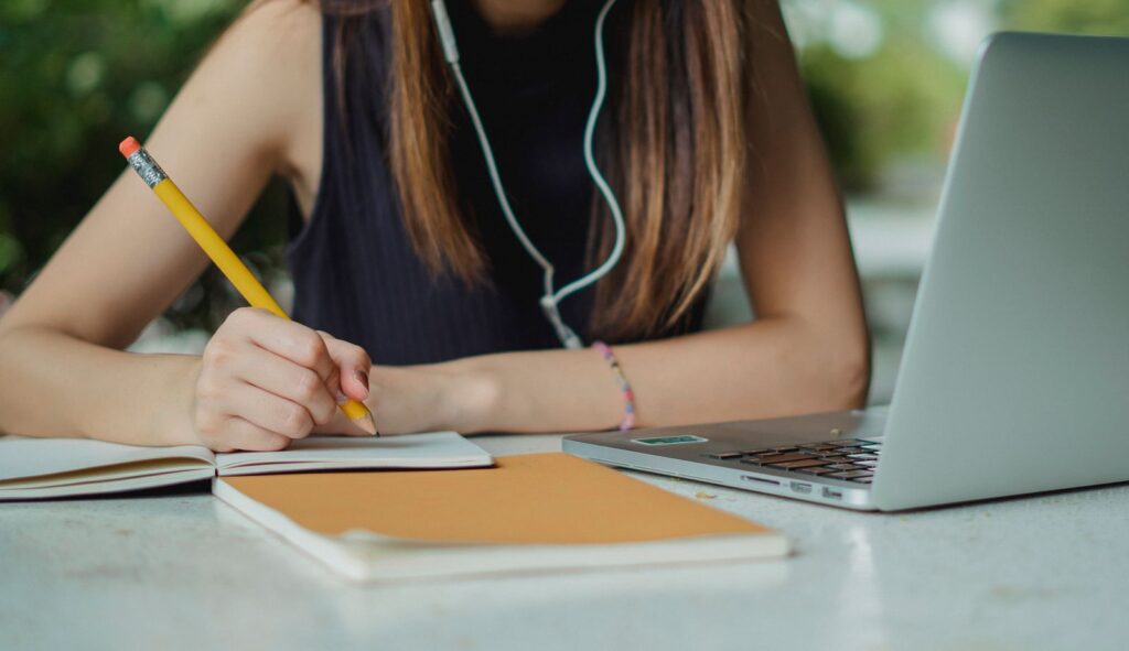 Student writing in a paper notebook with a laptop open nearby