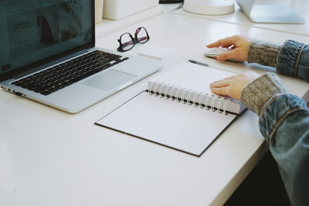 Person writing in a spiral notebook beside a laptop on a white desk